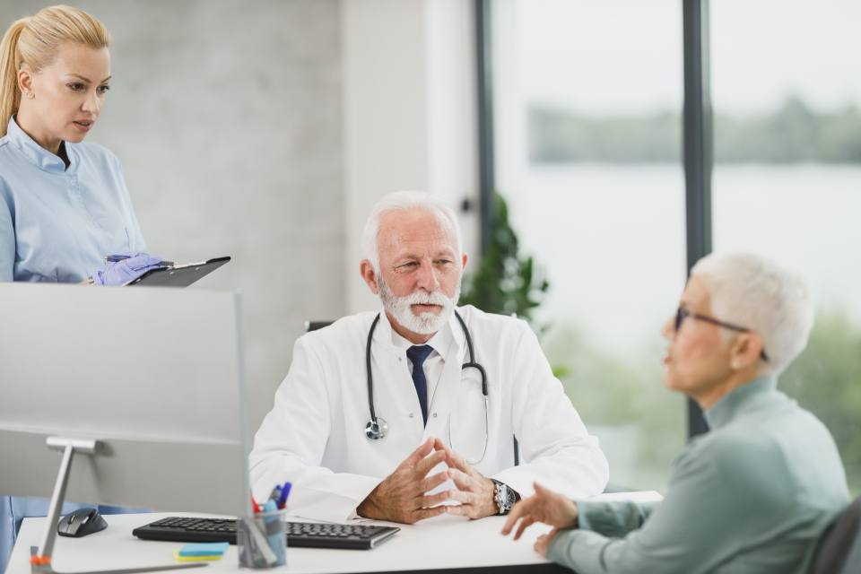 A doctor talks with an older patient across his desk while a nurse looks on